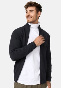 Pull zip noir foncé avec des poignets et un col côtelés, porté par-dessus un col roulé blanc. Texture lisse et design ajusté.