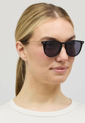 Frau mit blondem Haar, das zu einem niedrigen Pferdeschwanz gebunden ist, trägt schwarz gerahmte Sonnenbrillen mit goldenen Bügeln und ein weißes T-Shirt mit Rundhalsausschnitt vor einem schlichten Hintergrund.