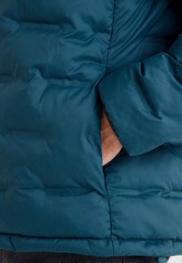 Teal puffer jacket with quilted texture, featuring a side pocket and elastic cuffs. Material is smooth and water-resistant.