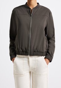 Svart bomberjacka med dragkedja fram, elastiska mudd och nederkant, slät tyg och utan krage. Buren över ljusa byxor.