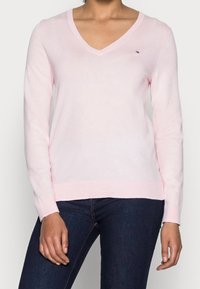 Jasnoróżowy sweter z dekoltem w serek, wykonany z miękkiej dzianiny, z długimi rękawami i ściągaczami, z małym emblematem logo na piersi.