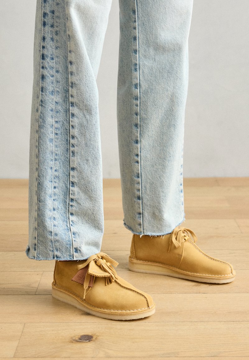 Geel suède enkelboots met franjes, platte zolen en gestikte accenten, gecombineerd met lichtblauwe denim jeans. Houten vloer op de achtergrond.