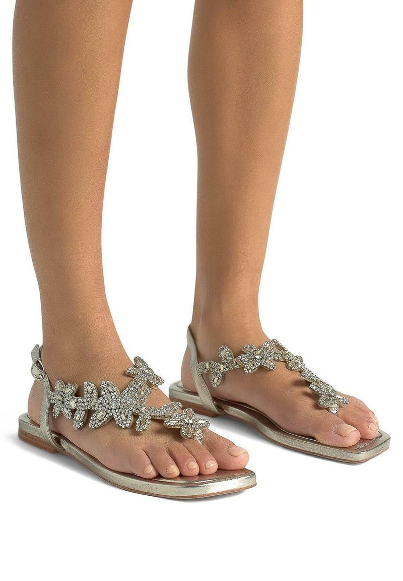 Sandali piatti argento con un design floreale arricchiti da pietre di strass trasparenti. Le cinghie sono eleganti e la suola ha un profilo piatto con una larghezza ridotta.