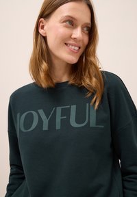 Mörkgrön sweatshirt med "JOYFUL" tryckt i stora bokstäver. Tillverkad av mjukt tyg, med rund nacke och avslappnad passform.