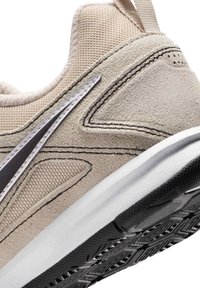 Κοντινό πλάνο μπεζ αθλητικού παπουτσιού με υφές από σουέτ και δίχτυ, μαύρη και λευκή σόλα, και μερικώς ορατή μαύρη και λευκή λεπτομέρεια λογότυπου.