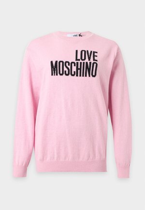 Жена, носеща розов пуловер с текст "LOVE MOSCHINO", черни панталони с бял сърцевиден мотив и черни обувки с отворени пръсти и панделки.
