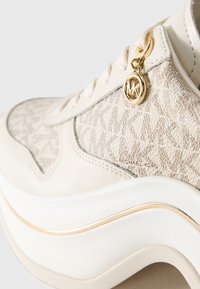 Prim-plan al unor sneakeri bej Michael Kors cu material texturat, talpă albă și charm cu logo-ul aurit MK lângă șireturi.