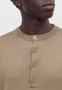 Gros plan d'une personne portant un pull marron clair à col rond avec quatre boutons marron clair sur le devant.