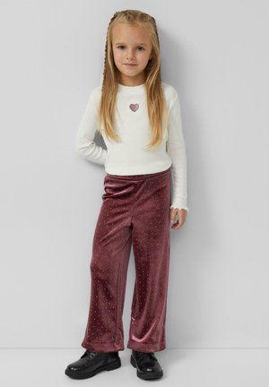 Ragazza giovane con capelli biondi lunghi che indossa un maglione bianco con cuore, pantaloni di velluto color bordeaux con pois, e stivali neri, in piedi contro un muro grigio.