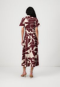 Robe maxi en bordeaux avec un motif géométrique crème, manches courtes, taille ceinturée et silhouette fluide. Portée avec des talons à lanières.