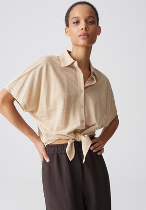 Femme portant une chemise beige à manches courtes nouée à la taille et un pantalon foncé avec taille élastique, debout les mains sur les hanches devant un fond blanc.