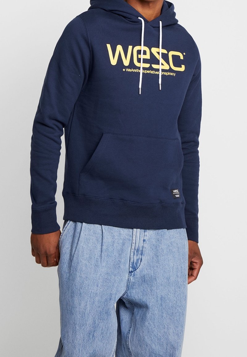 Sweatshirt à capuche bleu marine en matériau coton. Présente un grand logo jaune sur la poitrine et une poche kangourou. Corde blanche incluse.