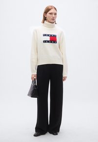 Pull à col roulé couleur crème avec un logo imprimé en bloc, associé à un pantalon large noir et un petit sac à main noir.