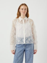 Blusa bianca trasparente con ricamo floreale, frontale con bottoni e colletto. Maniche lunghe con polsini a tinta unita in contrasto. Abbinata a jeans di denim.