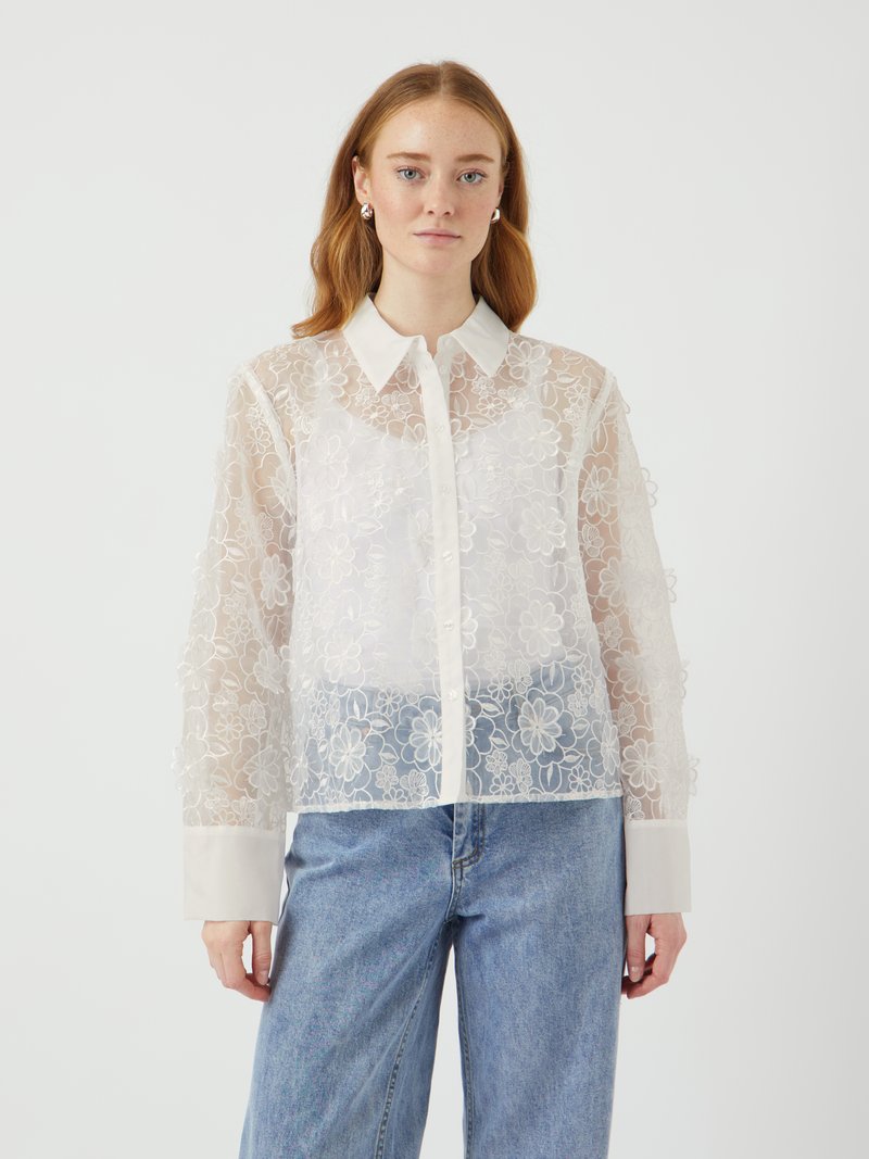 Blusa bianca trasparente con ricamo floreale, frontale con bottoni e colletto. Maniche lunghe con polsini a tinta unita in contrasto. Abbinata a jeans di denim.