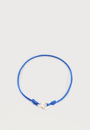 Bracelet en corde bleu réglable avec un anneau en argent et un fermoir à crochet sur un fond clair uni.