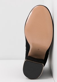 Scarpa nera in suede con tacco alto, che mostra una suola beige liscia, una suola in gomma nera testurizzata e una capocchia del tacco con dettagli dorati. Design pulito e minimalista.