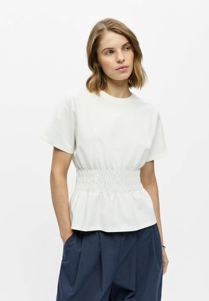 Vrouw met kort blond haar, die een wit kort mouwtje draagt met elastische taille en loszittende donkerblauwe broek, staand met handen in de zakken.