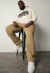 Pantalones cargo beige, sudadera crema con "SOHO NEW YORK" en letras negras en negrita, zapatillas blancas, sentado en un taburete de metal negro.
