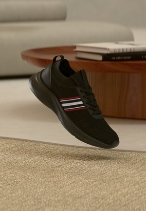 Fekete hálós sportcipő piros, fehér és kék csíkokkal, egy texturált bézs szőnyeg felett lebeg, egy fa dohányzóasztal előtt, melyen egymásra rakott könyvek vannak.