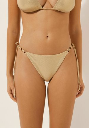 Bikini beige con tela texturizada, top de triángulo y braguitas con lazos laterales y detalles en forma de anillo, en el torso de una mujer con los brazos relajados.
