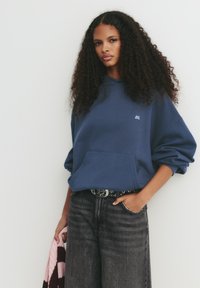 Mörkblå oversized hoodie med ficka fram och logotyp. Kombinerat med mörkgrå högmidjade jeans och ett mönstrat bälte.