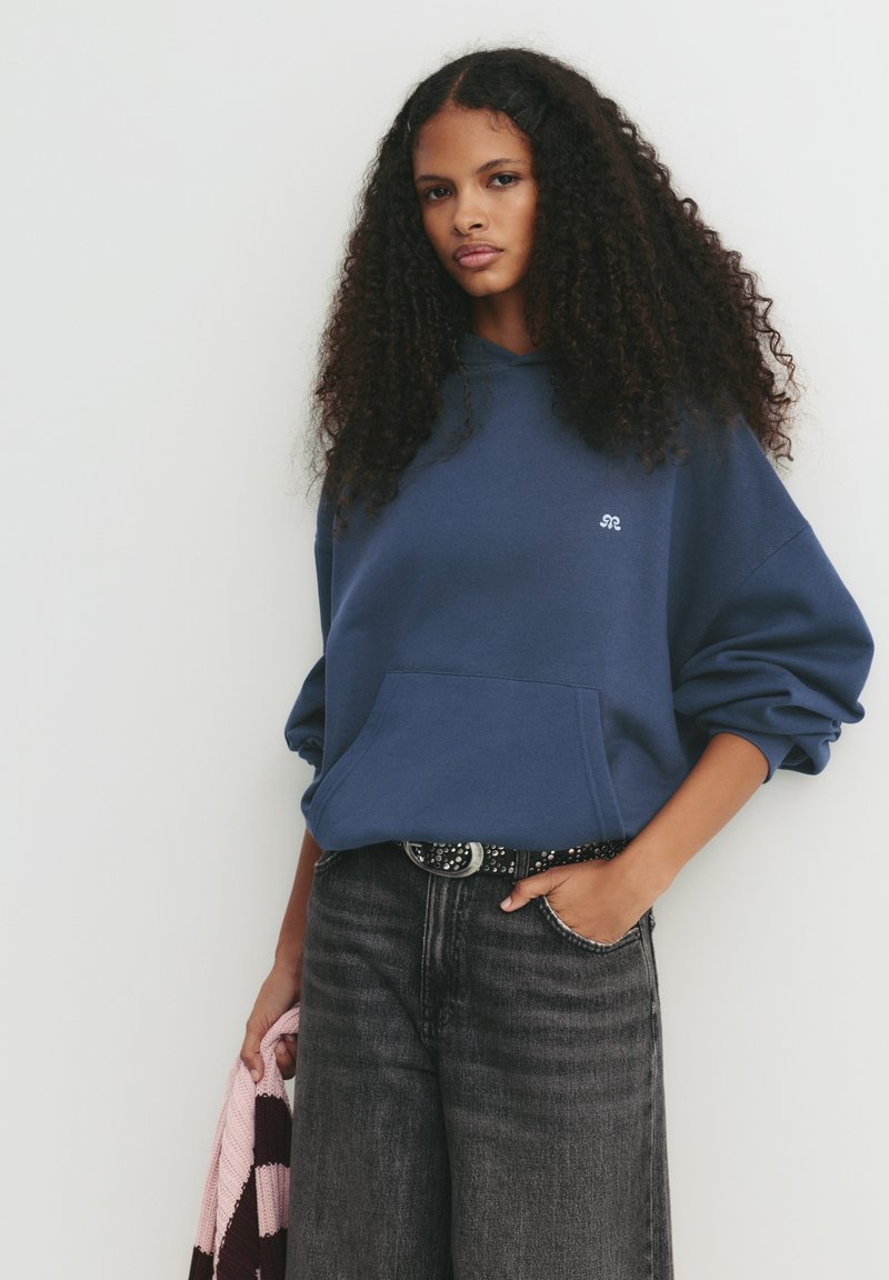 Mörkblå oversized hoodie med ficka fram och logotyp. Kombinerat med mörkgrå högmidjade jeans och ett mönstrat bälte.