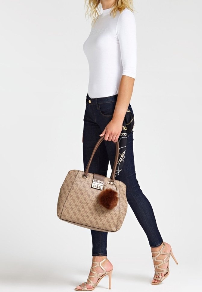Femme en haut blanc et jean foncé marchant, tenant un sac à main beige à motif avec un accessoire pompon, portant des sandales à talons hauts couleur nude.