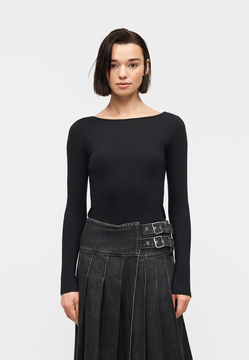 Femme aux cheveux courts et foncés portant un haut noir à manches longues côtelé et une jupe plissée sombre avec deux détails de boucle.