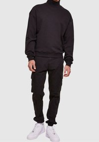 Sudadera negra de cuello alto con hombros caídos, combinada con pantalones cargo negros con bolsillos y zapatillas blancas. Diseño simple y funcional.
