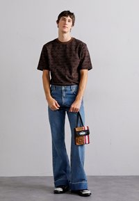 Tricou scurt cu maneci, de culoare maro și cu un model, asortat cu blugi evazați de culoare bleu deschis și o geantă mică maro, roșie și neagră. Pantofii negri completează ținuta.