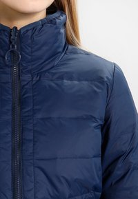 Tmavě modrá prošívaná bunda s vysokým límcem, předním zapínáním na zip a prošitým vzorem. Má kulatý přívěsek na zipu a je vyrobena z lehkého materiálu.