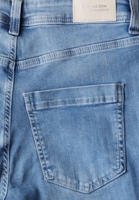 Pantalones vaqueros de mezclilla azul claro con un solo bolsillo trasero, costuras reforzadas y una etiqueta de tela en la cinturilla. Textura suave.