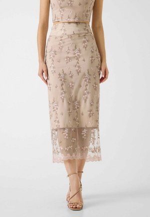 Beżowa, przezroczysta spódnica midi z haftem kwiatowym i podszewką, noszona z dopasowanym topem typu crop i beżowymi sandałami na wysokim obcasie z paskami.