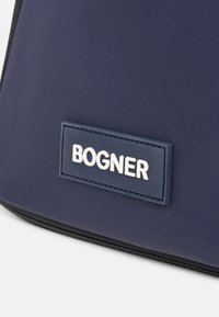 Marineblå nylon taske med et fremtrædende hvidt "BOGNER" logo på et tekstureret mærke, med en sort lynlås langs kanten.