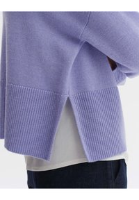 Maglione a maglia viola chiaro con orlo a coste e spacchi laterali, abbinato a una maglietta bianca e jeans blu scuro.