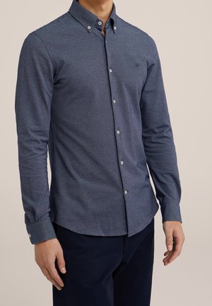 Homme portant une chemise bleue cintrée à manches longues avec col boutonné et pantalon foncé, debout devant un fond uni.