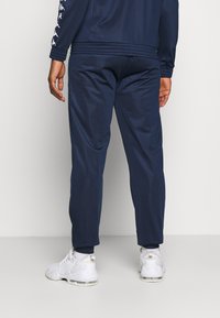 Sötétkék sportnadrág sima, szintetikus anyagból, szűkített lábakkal és bordázott mandzsettákkal, fehér sportcipővel párosítva.