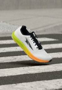 Λευκό και μαύρο αθλητικό παπούτσι με σόλα σε διαβάθμιση κίτρινου-πορτοκαλί, αιωρούμενο στον αέρα πάνω από διάβαση πεζών σε πόλη με διάσπαρτα σωματίδια βρωμιάς.