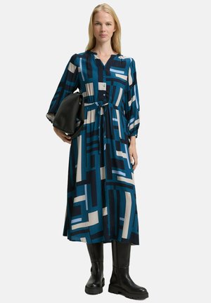 Langärmliges Hemdblusenkleid in einem geometrischen Muster aus Blau, Schwarz und Creme; der Stoff wirkt weich mit einer taillierten Taille; getragen mit schwarzen Stiefeln.