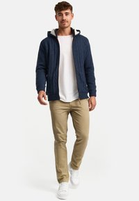 Navy knitted hooded sweater with a cable pattern, paired with a white t-shirt and beige pants. White sneakers complete the outfit.