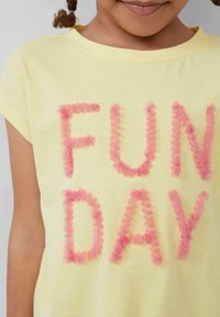 Żółta bawełniana koszulka z krótkim rękawem, z okrągłym dekoltem, na której widnieją słowa "FUN DAY" w puszystych różowych literach. Gładka tekstura.