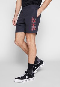 Shorts de bain bleus marine avec taille élastique, présentant un imprimé graphique rouge et teal sur le côté. Associés à des baskets noires et des chaussettes à motif.