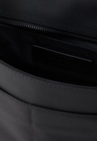 Εσωτερικό μαύρης υφασμάτινης τσάντας με φερμουάρ, που διαθέτει λείο, ματ λογότυπο και υφασμένη επένδυση για επιπλέον δομή.