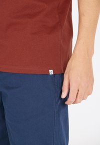 Marinröd t-shirt i bomull med korta ärmar, som har en liten vit märkestag på den nedre fållen, tillsammans med marinblå shorts.