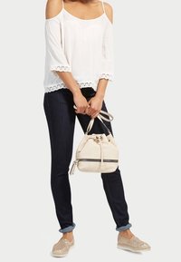 Biała bluzka z odkrytymi ramionami z koronkową lamówką, ciemne dżinsy i złote buty wsuwane. Trzyma beżową torbę typu bucket z czarnymi akcentami i paskiem na ramię.