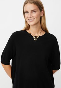 Smilende kvinde med langt blond hår bærer en sort kortærmet top og en guldkæde med flere bladformede vedhæng.