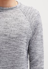 Suéter gris con un patrón de tejido texturizado y motas de gris más oscuro. El cuello redondo y las mangas raglán ofrecen un ajuste relajado.