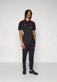 Čierny športový tréningový oblek s červenými doplnkami. Tričko má golier a logá; nohavice majú biele bočné pruhy a priliehavé manžety.