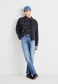 Chaqueta de mezclilla negra con botones plateados, camiseta gris, jeans azul claro y una gorra azul claro, combinados con zapatos negros.